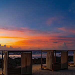 Wooden beach cabanas with flowing white curtains on sandy shore at vibrant sunset overlooking ocean